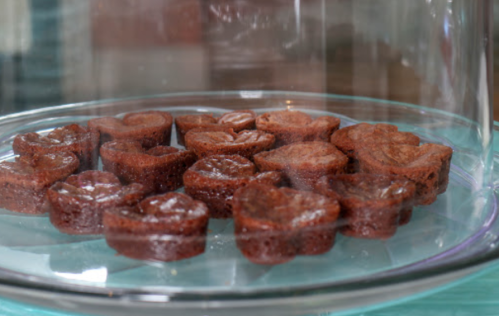 A glass cake dome displaying heart-shaped chocolate treats on a turquoise surface.