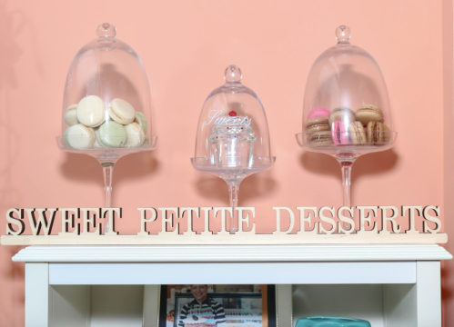 Three glass cloches display colorful macarons on a shelf with a sign that reads "Sweet Petite Desserts."