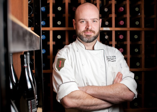 A chef in a white coat stands confidently with arms crossed in a wine cellar, surrounded by wine bottles.