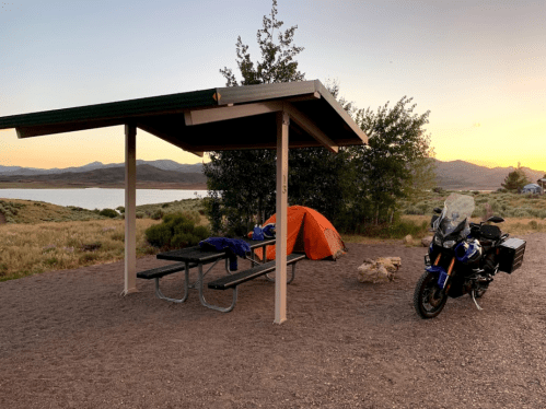 A campsite with a picnic table, orange tent, and motorcycle at sunset near a lake and mountains.