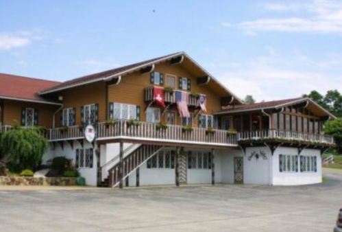 A large, two-story wooden building with balconies, flags, and a landscaped entrance on a sunny day.