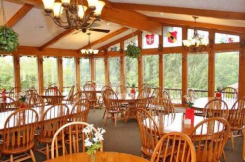 Bright dining area with wooden tables and chairs, large windows, and greenery, offering a scenic view.