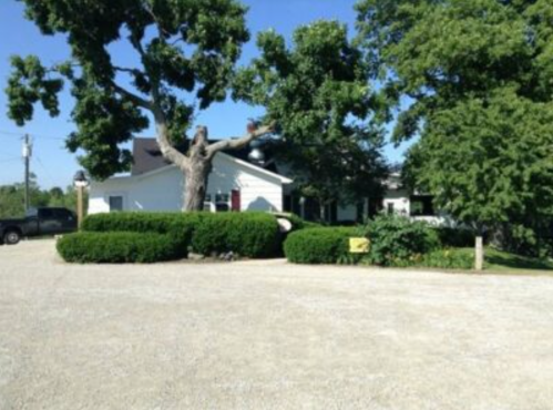 A gravel driveway leads to a white house surrounded by green bushes and trees under a clear blue sky.