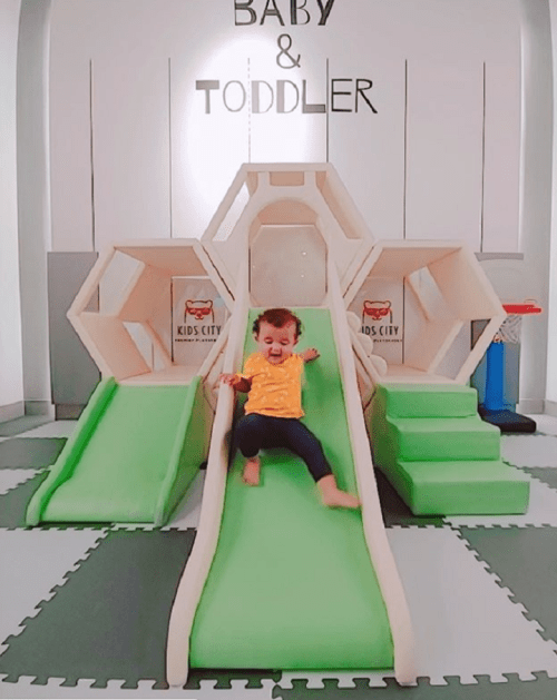 A toddler in a yellow shirt joyfully slides down a green slide in a play area designed for babies and toddlers.