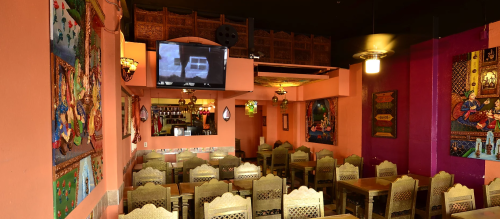 Interior of a vibrant restaurant with wooden tables, ornate chairs, colorful wall art, and a TV on the wall.