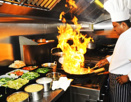 A chef stirs a pan over a flame in a busy kitchen, surrounded by fresh ingredients and cooking equipment.