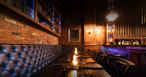 Cozy restaurant interior with brick walls, dim lighting, and a long table set with candles. Bookshelves in the background.