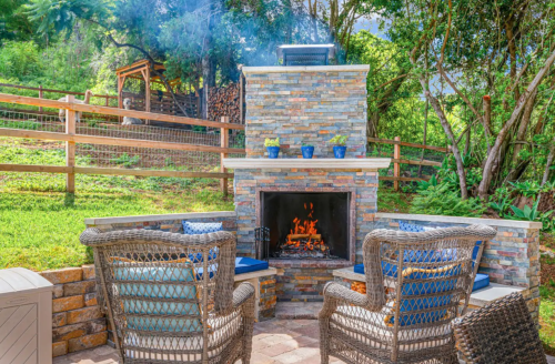 Cozy outdoor seating area with a stone fireplace, surrounded by greenery and wooden fencing.