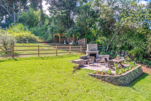 A landscaped backyard featuring a stone patio, seating area, and a fire pit, surrounded by greenery and a wooden fence.