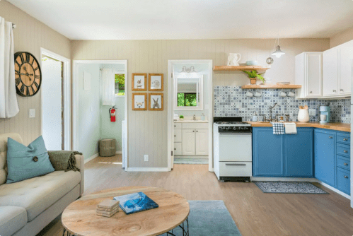 Bright, modern living space featuring a cozy sofa, a blue kitchen, and light wood flooring with decorative accents.