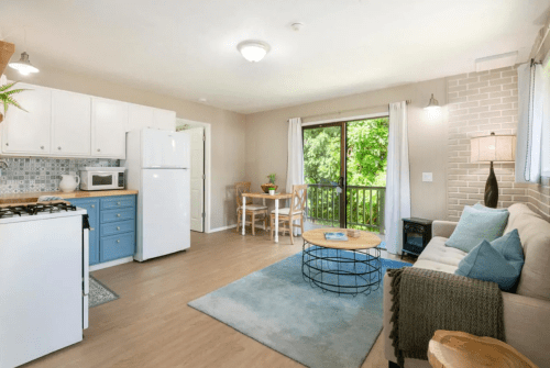 Bright, modern kitchen and living area with a sofa, dining table, and large window overlooking greenery.