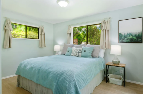 Cozy bedroom with a light blue quilt, two windows, and soft lighting from bedside lamps. Greenery visible outside.