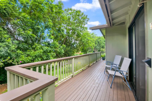 A wooden balcony with two chairs overlooks a lush green landscape under a blue sky.