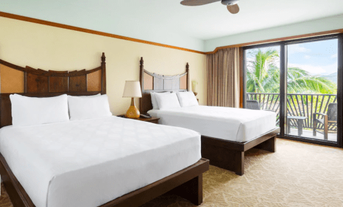 A bright hotel room with two queen beds, a lamp, and a balcony overlooking palm trees.