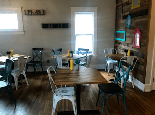 Cozy restaurant interior with wooden tables, colorful chairs, and a rustic wall, featuring bright windows and condiments.
