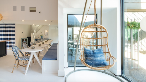 A bright, modern interior featuring a hanging chair, dining area, and large windows with a view of an outdoor space.