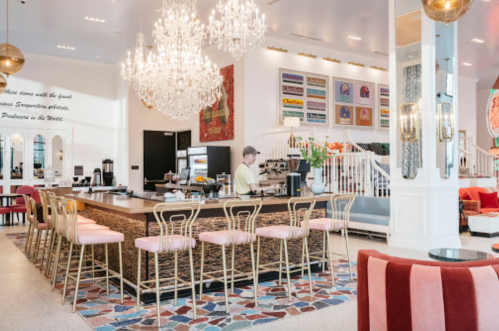 A stylish café with a chandelier, bar seating, and colorful decor, featuring a barista preparing drinks.