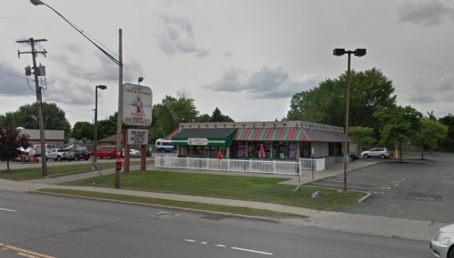 A restaurant with a green and red awning, outdoor seating, and a sign, located on a busy street with parked cars.
