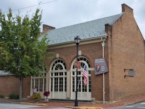 Historic brick building with large windows, a lamppost, and an American flag outside, surrounded by greenery.