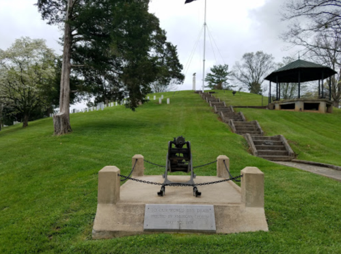 A cannon on display with a plaque, set on a grassy hill with steps leading to a gazebo and trees in the background.