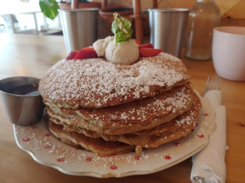 A stack of fluffy pancakes topped with strawberries, whipped cream, and powdered sugar, served with syrup on the side.