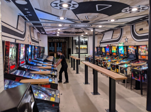A spacious arcade with multiple pinball machines and a person playing, featuring a stylish mural on the ceiling.