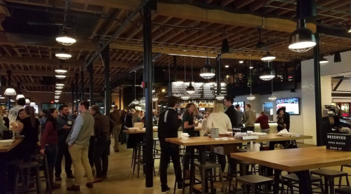 A lively restaurant interior with people socializing, tables, and a bar area, featuring warm lighting and a modern design.