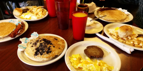 A table with several plates of breakfast food, including pancakes, eggs, bacon, and fruit, with drinks in red cups.