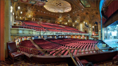 A panoramic view of a grand theater interior, featuring red seats and ornate architectural details.