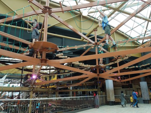 Two children navigate a rope course suspended above a colorful indoor play area with various structures.