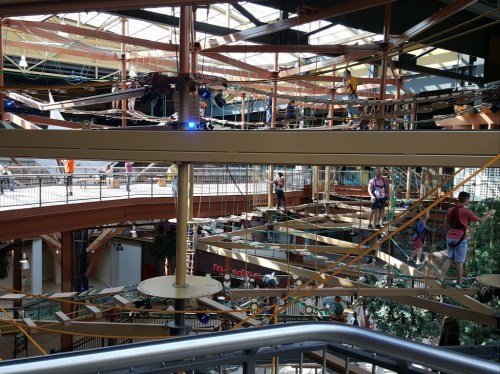 A multi-level indoor ropes course with people navigating obstacles high above a shopping area.