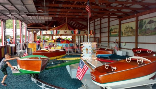 A vintage amusement park ride featuring wooden boats, colorful flags, and a carousel in the background.
