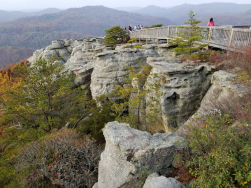 A scenic overlook with a wooden walkway, surrounded by rocky cliffs and autumn foliage in the background.