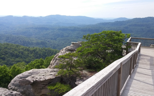 A scenic view from a wooden overlook, showcasing lush green mountains and a clear sky in the distance.