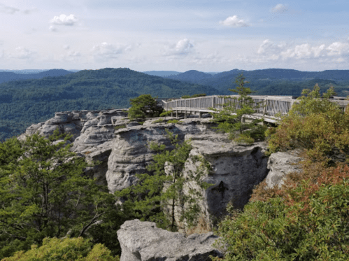 A scenic view from a rocky overlook, featuring lush green trees and distant mountains under a partly cloudy sky.