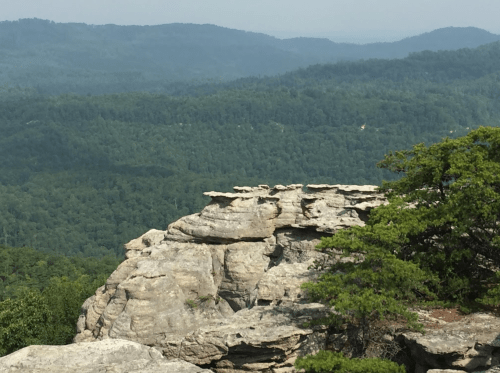 A rocky cliff overlooks a lush green valley and distant mountains under a hazy sky.