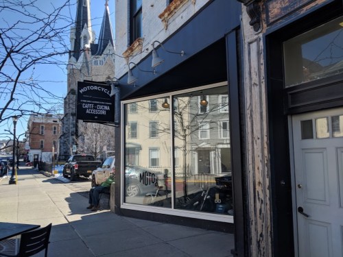 A street view of a café with large windows, featuring a motorcycle, and a church spire in the background.