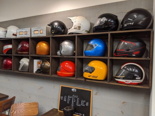 A wooden shelf displaying various motorcycle helmets in different colors and styles, with a chalkboard sign below.