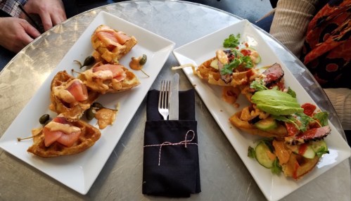 Two plates of waffle tacos topped with various ingredients, served on a round table with a fork and napkin.