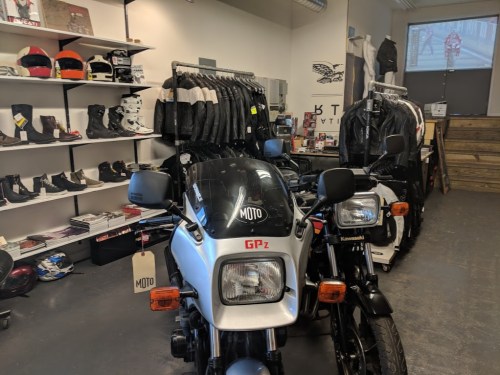 A motorcycle shop featuring two bikes, riding gear, and accessories displayed on shelves and racks.