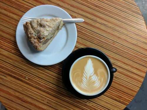 A slice of cake on a white plate next to a black cup of coffee with latte art, on a wooden table.