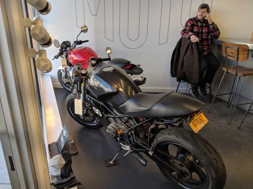 A black motorcycle and a red motorcycle are parked inside a cafe, with a man sitting nearby in a plaid shirt.