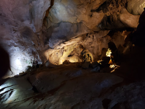 A dimly lit cave interior with rocky formations and shadows, illuminated by soft lights in various areas.