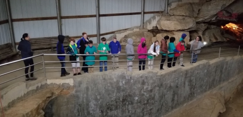 A group of children in colorful jackets stands along a railing inside a cave, observing their surroundings.