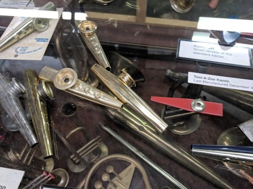 A display case featuring various kazoos, including a small version and a Tool-A-Zoo kazoo, among other musical instruments.
