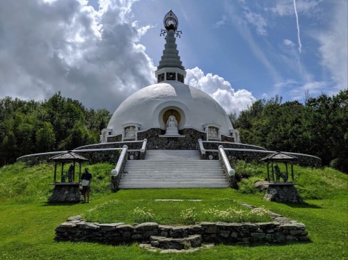 A white stupa surrounded by greenery under a blue sky with clouds, featuring a statue inside and stone pathways.