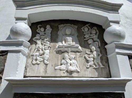 Relief sculpture depicting figures around a seated Buddha, framed by decorative elements on a stone wall.