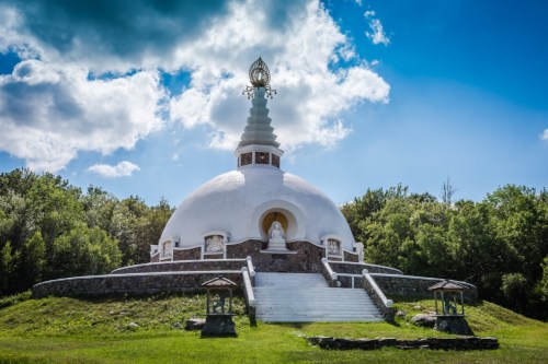 A serene white stupa surrounded by greenery under a blue sky with fluffy clouds.