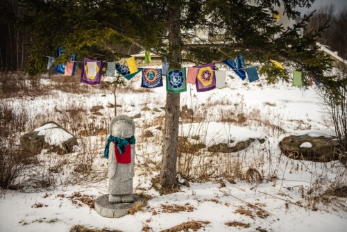 A stone statue stands under a tree, surrounded by colorful prayer flags in a snowy landscape.