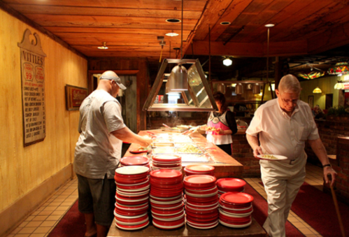 A buffet with diners serving food, surrounded by stacked red plates in a rustic restaurant setting.